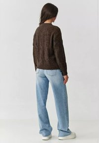 Brązowy sweter z dzianiny o teksturowanym wzorze, w połączeniu z jasnoniebieskimi spodniami o szerokich nogawkach i białymi sneakersami, widziany z tyłu.