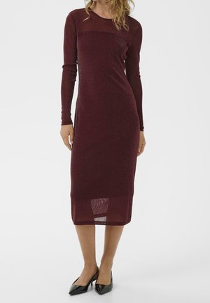 Kobieta w długim rękawie, dopasowanej sukience do kolan w kolorze burgundowym z subtelnym połyskiem oraz czarnych szpilkach z ostrym noskiem.