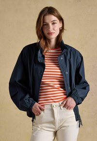 Kobieta ubrana w granatową kurtkę na czerwono-białej pasiastej koszulce, w kremowych spodniach, pozująca na beżowym tle.