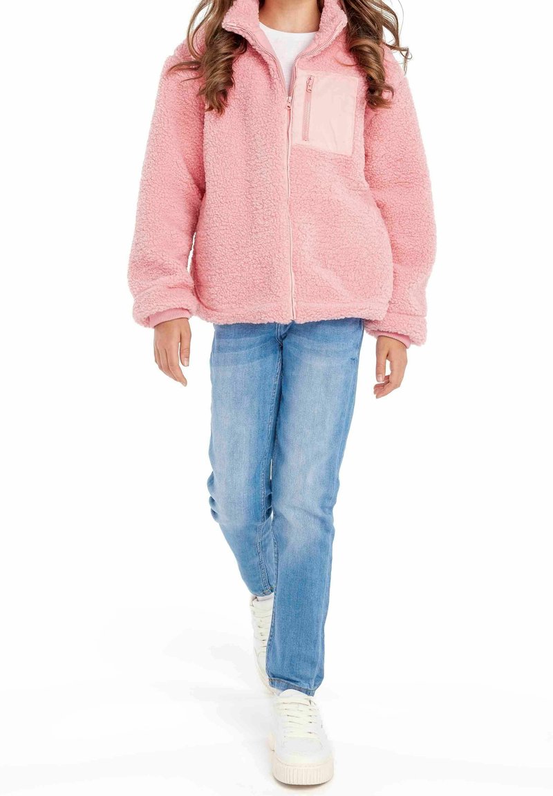 Personne portant une veste polaire rose zippée, un jean bleu clair et des baskets blanches, marchant vers l'avant sur un fond blanc.