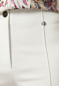 Pantaloni bianchi su misura realizzati in tessuto liscio, con una vestibilità elegante e un dettaglio in bottone argentato sulla vita. Blusa floreale testurizzata parzialmente visibile.