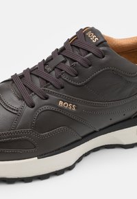 Sötétbarna bőr sneaker, kontrasztos bézs gumitalppal, kerek orral, puha belsőrésszel és a "BOSS" logó domborításával az oldalon.