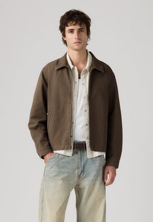 Ung mand iført brun jakke over beige ternet skjorte og hvid tanktop, kombineret med lyseblå jeans, stående mod grå baggrund.