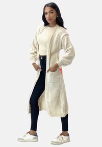 Cardigan lungo beige con polsini a coste, apertura frontale e due tasche laterali. Indossato sopra un maglione chiaro e jeans skinny neri. Le sneakers sono bianche con accenti grigi.