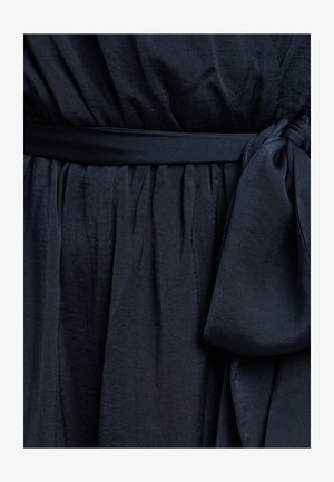 Marineblauwe jurk met een gestructureerde stof, een geraffineerde taille en een gestrikt riempje. De glans van de stof zorgt voor een subtiele glanzende uitstraling.