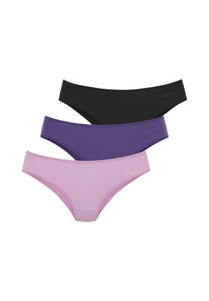 Donna con capelli ricci in piedi a piedi nudi indossa un reggiseno in pizzo viola e slip abbinati su uno sfondo chiaro uniforme.