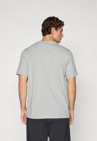 Mann mit lockigem Haar, der von hinten schaut, trägt ein schlichtes, hellgraues T-Shirt mit kurzen Ärmeln und dunkle Hosen vor einem einfarbigen Hintergrund.