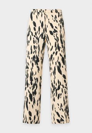 Femme aux longs cheveux foncés porte un costume beige à pois noirs avec pantalon assorti, un haut noir transparent et des chaussures plates noires, debout de profil.