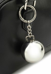 Schwarze Lederhandtasche mit einem glänzenden silbernen Kugelanhänger, der an einer metallischen Kette und einem Ring befestigt ist. Strukturierte Oberflächendetails sichtbar.