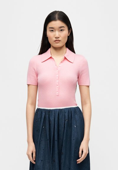 Νεαρή γυναίκα με μακριά σκούρα μαλλιά φοράει ροζ κοντομάνικο πουκάμισο με κουμπιά και ψηλόμεση σκουρό μπλε φούστα με διακριτικές διακοσμήσεις.