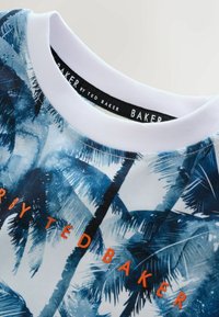 T-shirt à manches courtes avec un imprimé feuilles de palmier bleu vif, col blanc et texte orange "by TED BAKER" à l'avant. Texture de tissu lisse.