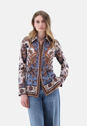 Femme aux cheveux blonds lâches portant une chemise à manches longues à motif floral bleu, marron et blanc, associée à un jean bleu clair.