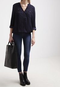 Svart knapp-up långärmad blus med två bröstfickor. Bärs med mörkblå skinny jeans och svarta klackade ankelstövlar, bärande en svart tote-väska.