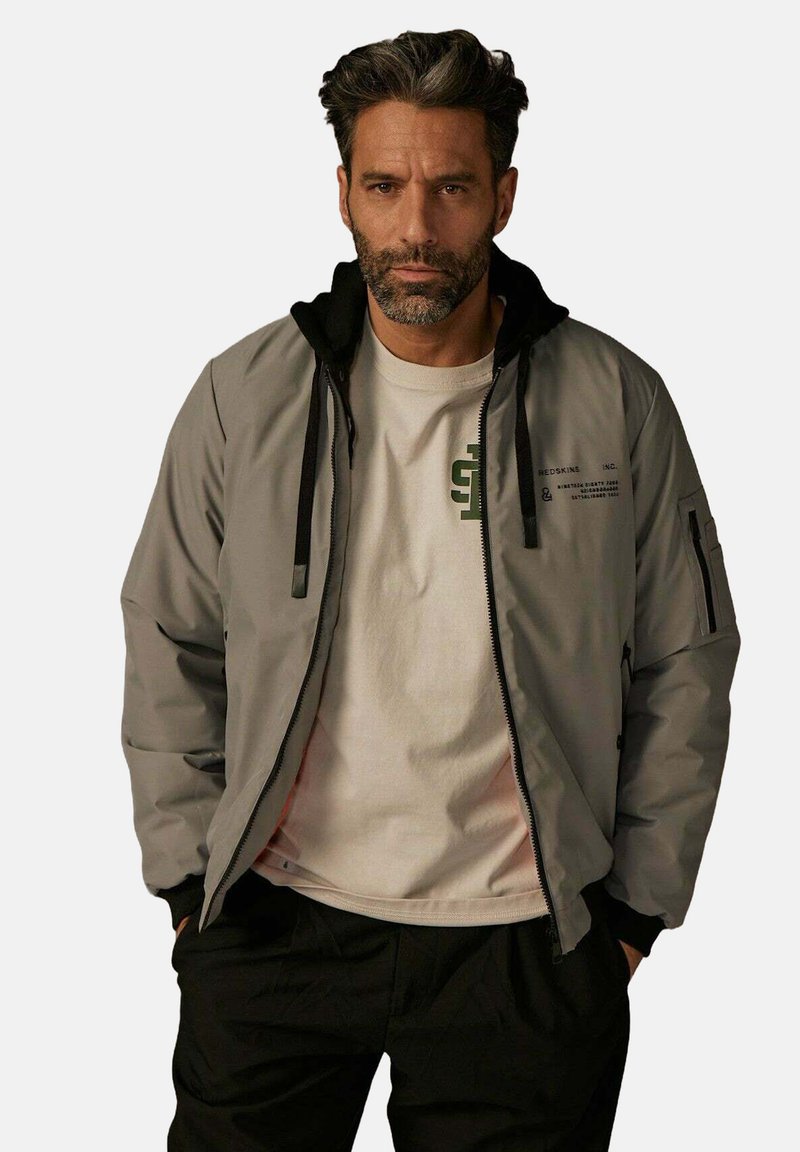 Veste grise avec une capuche noire, comportant des zips et des poches, superposée à un t-shirt de couleur claire avec du texte imprimé et des motifs.