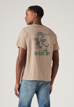 Beige katoenen t-shirt met een afbeelding van een kat in een hoed op ski's, met de tekst "GEEN DAGEN VRIJ" eronder in donkergroene print.
