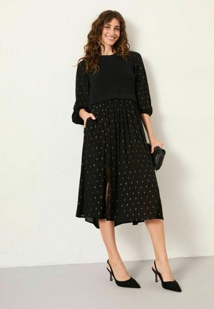 Schwarzes Kleid mit einem strukturierten Strickoberteil und einem fließenden, gemusterten Rock mit goldenen Akzenten. Lange Ärmel und seitliche Taschen sind enthalten.