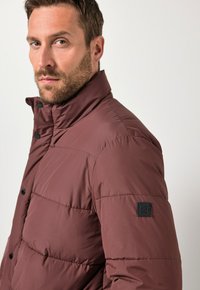 Bourgogne pufferjakke med høj krave, tekstureret stof, frontknapper og et kvadratisk sort logo patch på venstre ærme.