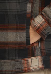 Flanellesärk, millel on musta, halli ja oranži ruuduline muster. Vasakul küljel on tasku ja nööpiga mansett. Kangas paistab pehme.
