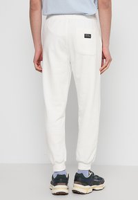 Witte trainingsbroek met een taps toelopende vorm, elastische tailleband en één achterzak. De stof lijkt zacht met een gladde textuur.