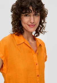 Camicia di lino arancione con maniche corte, colletto e chiusura con bottoni. Presenta un tessuto testurizzato e bottoni a contrasto.