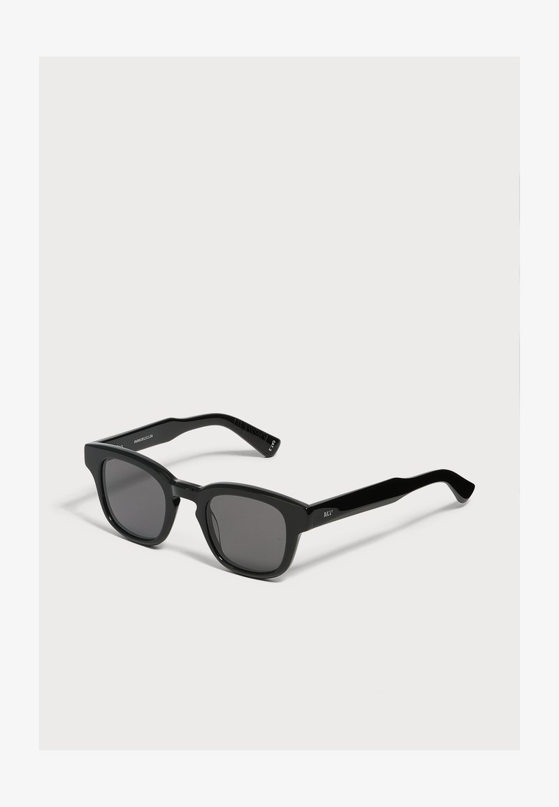 Lunettes de soleil à monture carrée noire avec des verres teintés foncés posées sur un fond gris clair.