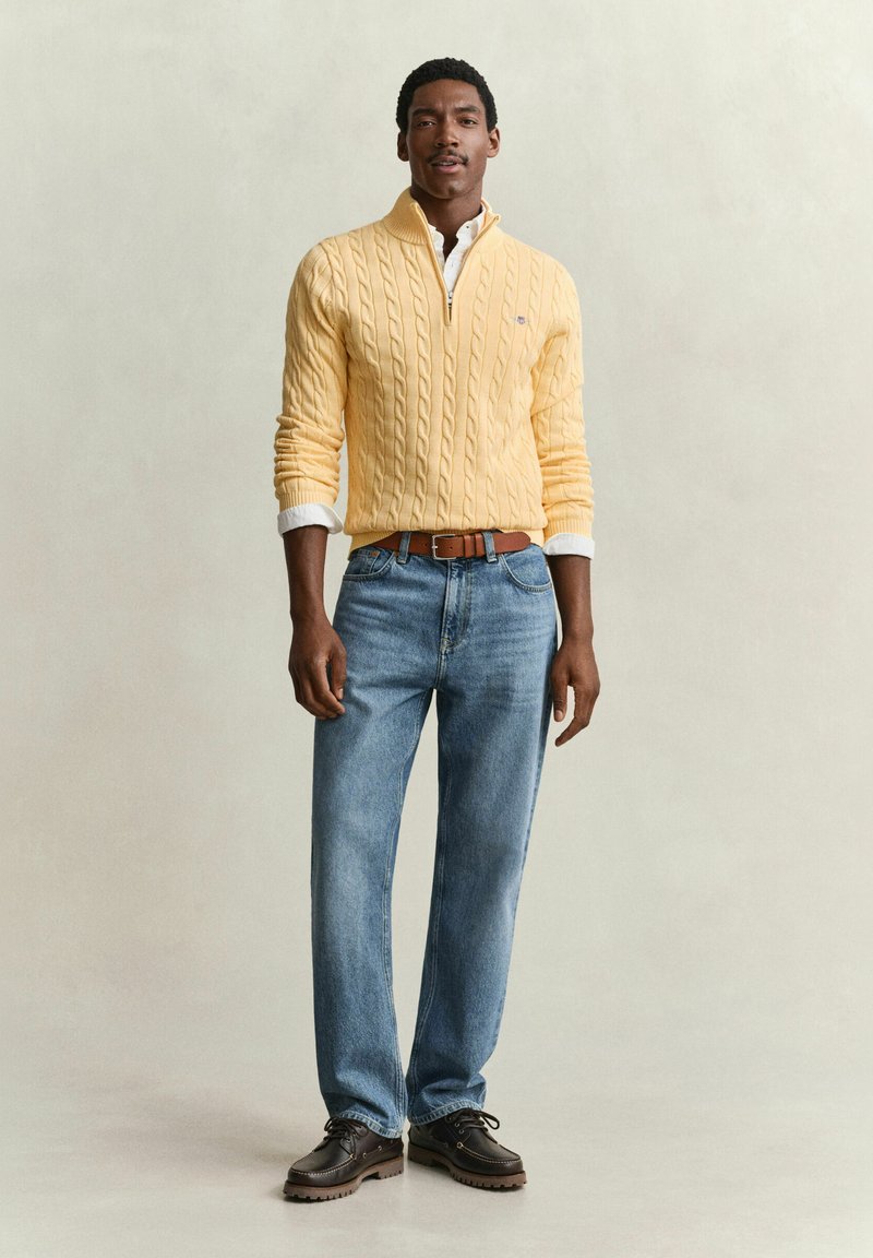 Man wearing yellow cable knit quarter-zip sweater, white shirt, light blue jeans, brown belt, and dark brown shoes standing against plain background.