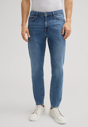 Light blue denim jeans with a slim fit, five-pocket design, and a button closure. Paired with white sneakers for a casual look.