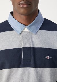 Tengerészkék, szürke és világoskék csíkos pólóing. Pamut anyagból, chambray gallérral és logóval a mellkason. Háromgombos sliccel.