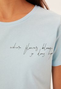 Camiseta de algodón azul claro con cuello redondo y mangas cortas. Presenta texto manuscrito en negro: "donde florecen las flores, allí también florece la esperanza."