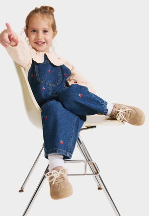 Young girl sitting cross-legged on a chair, smiling and pointing forward, wearing cherry-embroidered denim overalls and beige sneakers.