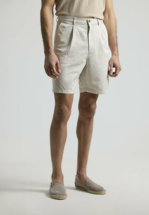 Pantalones cortos de algodón blancos con pliegues, con cierre de botón y bolsillos laterales, combinados con alpargatas de color gris claro. Diseño minimalista.