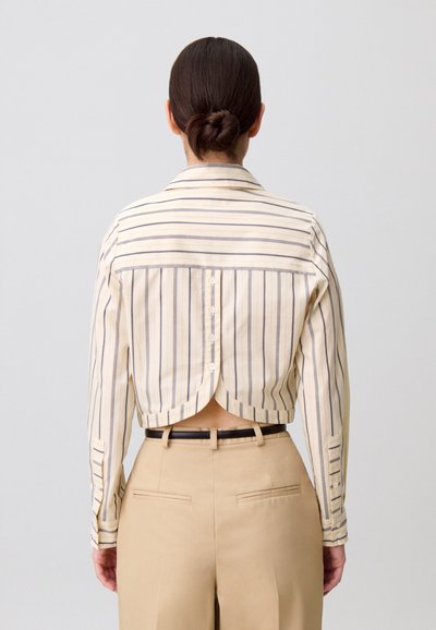 Camicia a righe color crema e blu navy con bottoni, design corto con orlo arrotondato, maniche lunghe e dettaglio posteriore con spacchi. Abbinata a pantaloni beige.