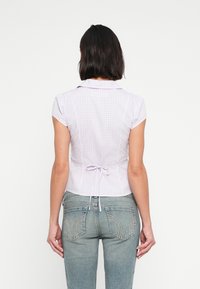 Μπλούζα με κοντά μανίκια, σε μοτίβο λιλά καρό γκίνχαμ, με γιακά, εφαρμοστή γραμμή και λεπτομέρεια δέσιμου στη μέση. Φορεμένη με ανοιχτόχρωμο τζιν.