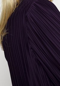 Lila plisserad tyg med vertikala linjer, som har en strukturerad yta. Materialet är slätt med en lätt glans.