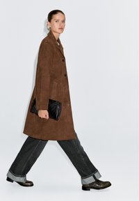 Bruine suède jas met een knoopsluiting, zijzakken en een losse pasvorm, gecombineerd met donkere jeans en zwarte leren enkellaarsjes.