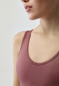 Sportovní podprsenka v růžové barvě vyrobená z hladkého a elastického materiálu; má kulatý výstřih a ploché švy na ramínkách.