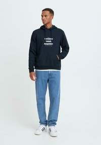 Navy hoodie with front pocket, featuring white text “EXPAND YOUR MINDSET,” paired with light blue jeans and white sneakers with black stripes.