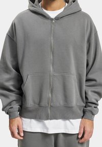 Sweat à capuche gris zippé avec cordon de serrage, grandes poches avant, poignets côtelés et texture douce, superposé sur un t-shirt blanc.