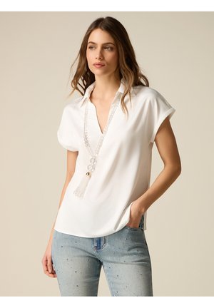 Donna con capelli castano lunghi che indossa una camicetta bianca a maniche corte, jeans blu con piccoli ornamenti e una lunga collana bianca di perline con nappe.