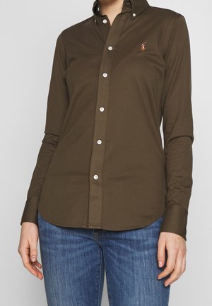 Kobieta w oliwkowozielonej koszuli na guziki z haftowanym logo polo gracza, w niebieskich dżinsach, stoi na jednolitym tle.