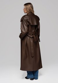 Brązowy płaszcz trench z sztucznej skóry z paskiem w talii, naramiennikami oraz szerokim kołnierzem. Płaszcz sięga do połowy łydki i ma gładką teksturę.