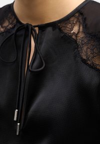 Blouse noire texturée avec panneaux en dentelle aux épaules et encolure à nouer avec ouverture en forme de goutte d'eau à l'avant, avec cordons à embouts métalliques.