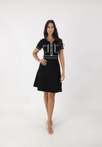 Schwarzes Kleid mit tailliertem Oberteil und ausgestelltem Rock, ausgestattet mit weißen Rändern, goldenen Knopfdetails und zwei vorderen Taschen. Klare Absatzschuhe.