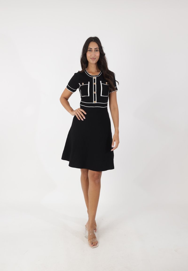 Schwarzes Kleid mit tailliertem Oberteil und ausgestelltem Rock, ausgestattet mit weißen Rändern, goldenen Knopfdetails und zwei vorderen Taschen. Klare Absatzschuhe.