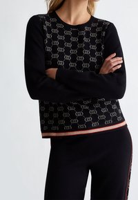 Femme portant un pull noir avec un motif de maillon en beige et un pantalon noir assorti avec des détails en rouge et beige. Bras croisés.