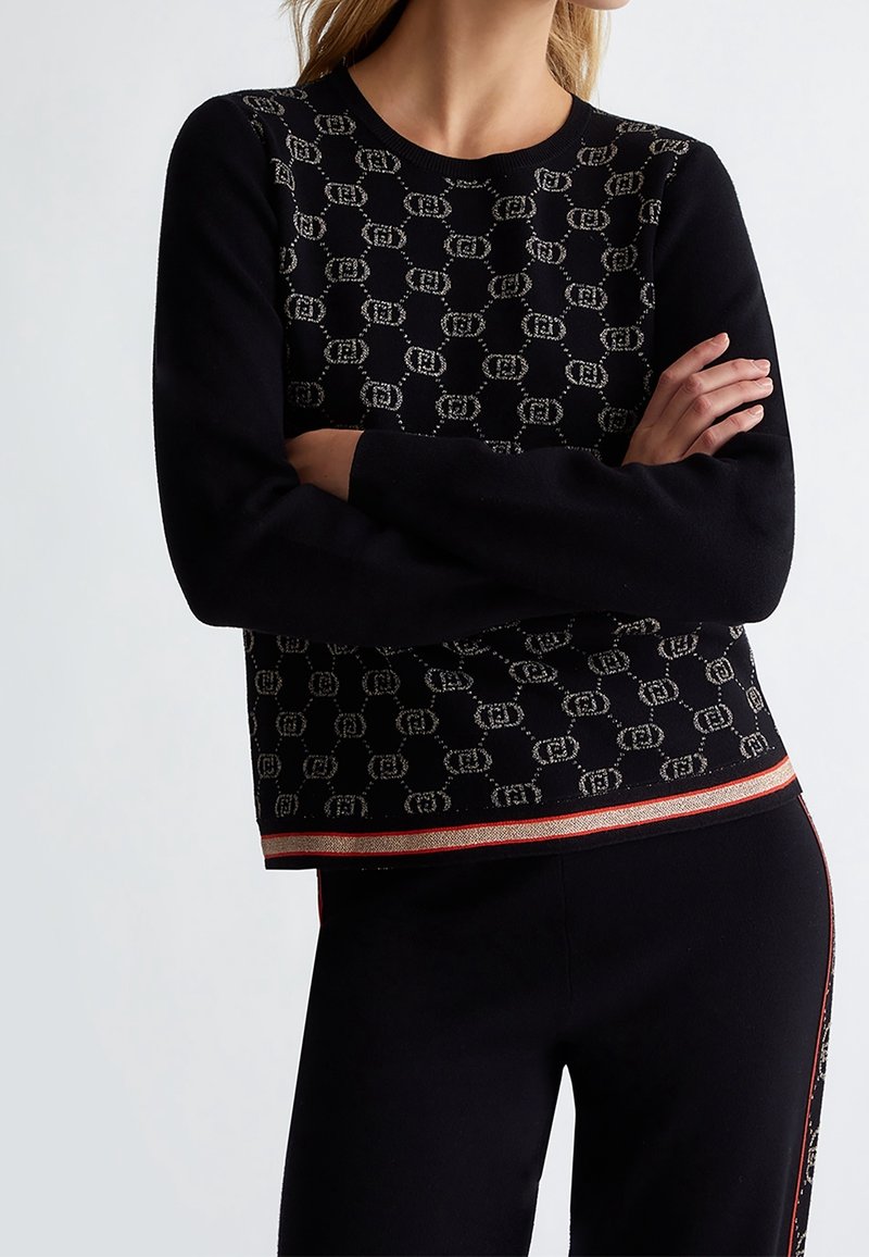 Femme portant un pull noir avec un motif de maillon en beige et un pantalon noir assorti avec des détails en rouge et beige. Bras croisés.