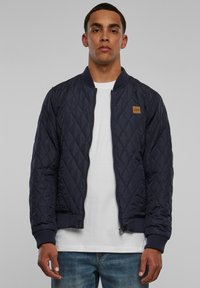 Námornícka prešívaná bomber bunda, ktorá disponuje zipsovým uzáverom, ribovanými manžetami a spodným okrajom a malým koženým logo nášivkom v béžovej farbe na hrudi. Pod ňou je biele tričko.