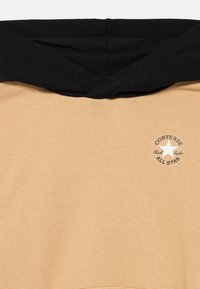 Μπεζ φούτερ με κουκούλα και μαύρο κορδόνι. Διαθέτει λευκό λογότυπο αστεριού με το κείμενο "Converse" στο στήθος. Μαλακή υφή.