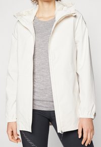 Femme portant une veste zippée écru avec des poignets élastiques, un t-shirt gris texturé et des leggings noirs sur un fond uni.