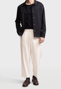 Zwarte button-up jas over een zwart shirt, gecombineerd met crèmekleurige plissébroek en bruine veterschoenen. Glad textuur en een relaxte pasvorm.